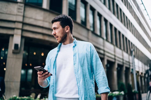 Pensive man with cellular device in hands thoughtful looking away during free time at urban setting, contemplative male blogger pondering on 4g internet connection tariff for smartphone gadget