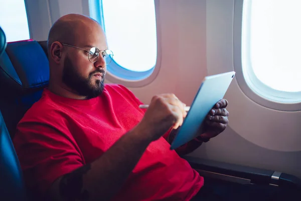 Caucasian man aircraft passenger touching tablet screen with digital pencil while sitting next to airplane window in cabin, Male enjoying his flight with wireless internet connection on board