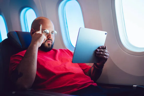 Young man flight passenger connecting to wireless internet on board with touch pad while sitting with pencil next to aircraft cabin window. Caucasian male working remote on freelance as digital nomad