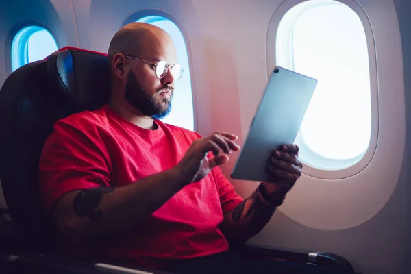 Caucasian man aircraft passenger touching tablet screen with hand while sitting next to airplane window in cabin, Male enjoying his flight with wireless internet connection on board