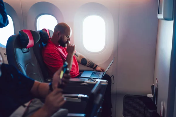 Young man flight passenger watching movie on board via entertainment Tv while sitting in comfortable airplane seat next to aircraft cabin window. Caucasian man enjoying travel via business class