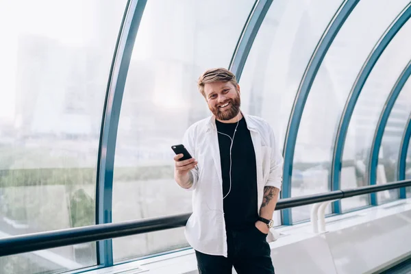 Half length portrait of happy bearded meloman in casual outfit smiling at camera while listening favourite audio podcast via electronic headphones, positive hipster guy enjoying music entertainment