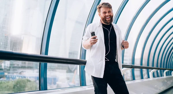 Half length portrait of positive hipster guy dancing at public place while listening music playlist via smartphone app and electronic earphones, funny male meloman enjoying sound record indoors