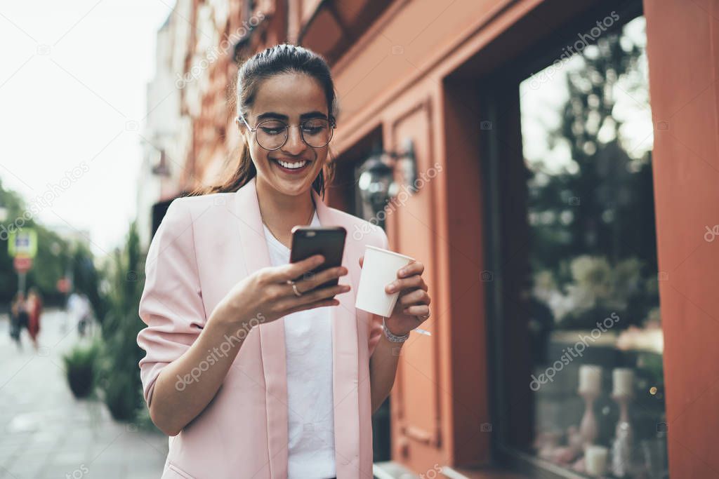 Cheerful woman in big classic glasses for provide eyes protection typing text message during time for exploring new city on vacations using public internet connection on modern cellphone gadget