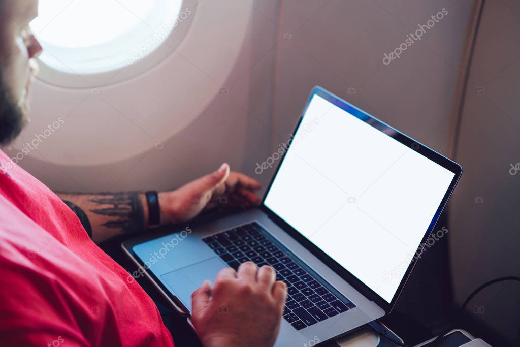 Young man flight passenger connecting to wireless internet on board with laptop while sitting next to aircraft cabin window. Mock up blank screen for web template, digital nomad work on freelance