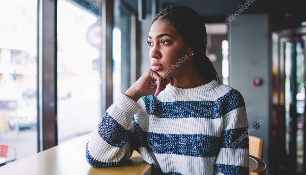 Dark skinned pensive hipster girl in trendy apparel thinking about life in comfortable university cafeteria, thoughtful female student pondering on upcoming school exams while spending time in campus