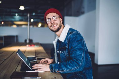Optik gözlüklü milenyum adamı, modern dokunmatik ped ile kahve dükkanında oturan ve diğer tarafa bakarken gülümseyen dijital tabletli, modaya uygun giyinen erkek blogcu