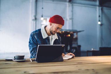 Web sayfalarında bilgi taramak ve ders kitabında genel kavramları kaydetmek için dokunmatik ped kullanan yakışıklı öğrenci, kahvede otururken yazar yazısı