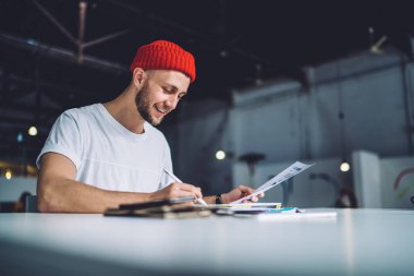 Eğilimli kırmızı şapkalı yakışıklı beyaz adam üniversite sınavlarına hazırlanmak için öğrenme süreci boyunca not tutuyor. Pozitif hipster çocuğu çalışma saatleri içinde SMS metni yaratıyor.