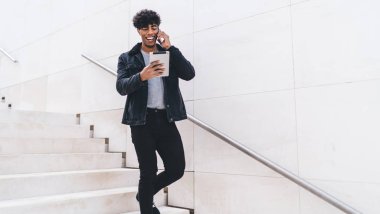 Moda kıyafetleri giymiş neşeli Latin adam komik içerikli e-posta alırken şaka tartışmak için arkadaşça bir telefon görüşmesi yapıyor, modern aletleri kullanan hippi adam dışarıda.