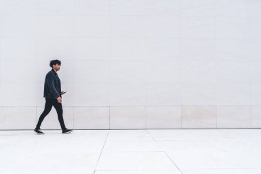 Modern dijital tabletli modaya uygun giyinmiş bir erkek öğrencinin, tanıtım amaçlı duvar arkasından şehir merkezine yürümesi, fotokopi alanı yakınlarındaki şık bir adamın reklam alanında buluşması.