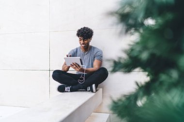 Elektronik kulaklık ve dokunmatik ped uygulaması ile eğlenceli müzik dinlemek için şehir merkezinde boş vakitlerin tadını çıkaran pozitif hipster adam, tabletle meloman arayan neşeli Latin adam web sitesi