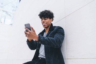 Cep telefonu cihazlı genç, olumlu hipster arkadaşı ile görüntülü iletişim kuruyor, mutlu milenyum erkek blogcusu akıllı telefon içeriğini okuyarak kentsel arka planda duruyor.