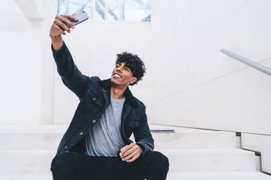 Hücresel aygıtlarla neşeli Latin bir adam akıllı telefon kamerasıyla selfie çekiyordu. Şehir merkezinde dinleniyordu. Sosyal ağlarda blog için video çeken mutlu genç bir erkek modern cep telefonu kullanıyordu.