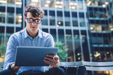Başarılı bir erkek tüccar internet sitelerinde finansal haberler okuyarak modern tablette 4G internet kullanarak modern gökdelenleri olan bir iş şehrinin caddesinde oturuyor.