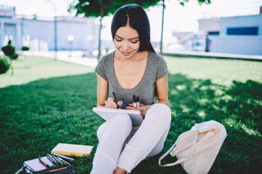 Şehir parkında boş zamanlarında üniversite dersleri için doğayla eskizler çizen pozitif kız öğrenci, tasarım okulunda eğitim için ders kitabını kullanarak sınava hazırlanan mutlu hippi kız.