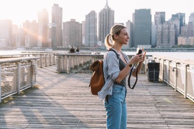Side view of positive Caucasian woman wanderer with old fashioned instant camera in hands looking around for taking few pictures of American view, concept of weekend getaway to Brooklyn district clipart