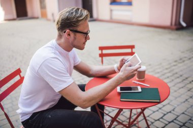 Millennial giyimli bir adam sokak kafeteryasında internet bağlantısı olan açık havada otururken dijital tablet kullanıyor, beyaz bir erkek internet sitesinden bilgileri kontrol ediyor.