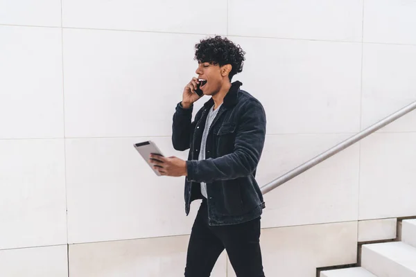 Shocked Latino man checking received email with account balance while walking at urban setting and making smartphone call for discussing information, hipster guy amazed from touch pad notification
