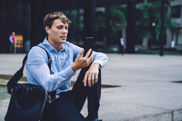 Caucasian businessman in formal wear making international cell conversation with partner while sitting at city area of financial district, experienced male trader in headphones watching video