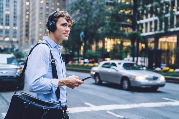 Successful male entrepreneur in formal wear holding modern smartphone device and using music application for listening audio via headphones for noise cancellation, Caucasian man in downtown