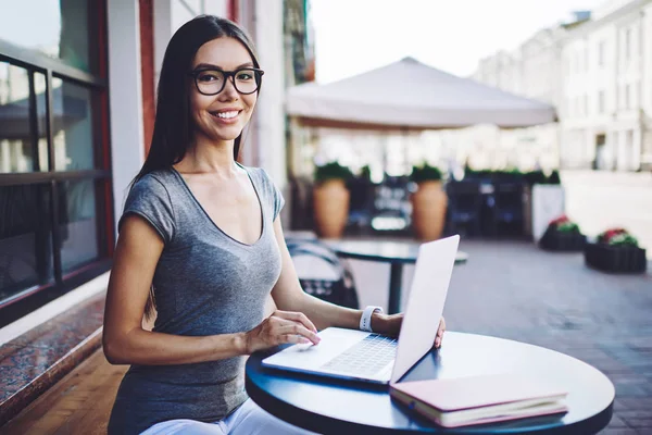 在街上的自助餐厅里 快乐的女自由撰稿人面带微笑 坐在笔记本电脑旁 戴着眼镜 享受着与现代上网本的远距离工作 — 图库照片