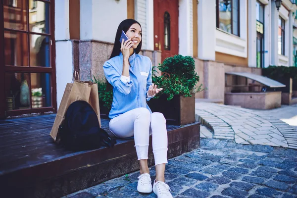 快乐的嬉皮士女孩在购物后通过手机应用程序与朋友交流 成功的女人带着背包和购物袋坐在城市里 打国际电话 — 图库照片