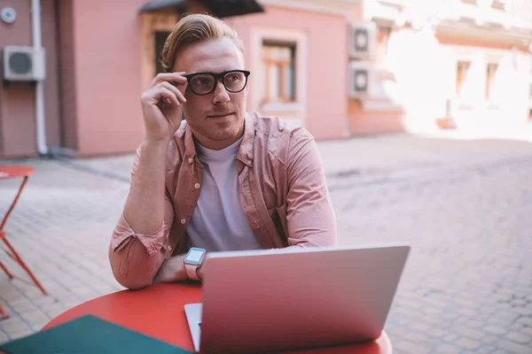 Contemplative hipster guy in classic glasses for vision protection sitting at cafe table and pondering on content for website, pensive hipster programmer working remotely with modern laptop computer