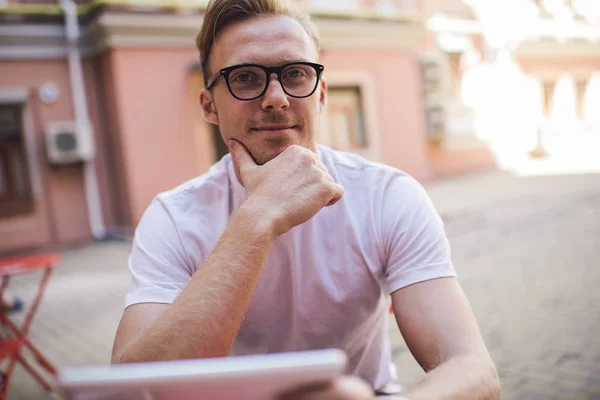 Portrait of Caucasian male blogger looking at camera while using digital tablet for messaging in web social networks, millennial young man pondering on app connection and online notification