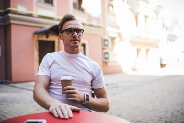 Thoughtful man in casual white t shirt looking away and thinking during coffee time in street cafeteria, stylish male student in optical eyewear holding takeaway cup with caffeine beverage