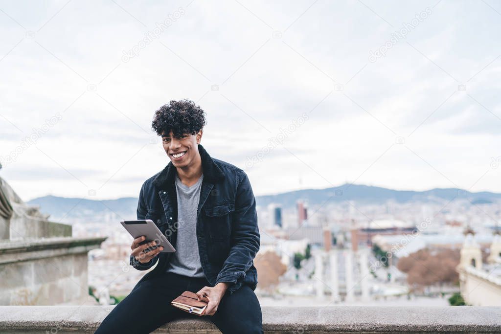 Portrait of happy cheerful male tourist dressed in trendy clothing resting in historic center of Spanish capital and smiling at camera during online chatting with followers from travel web blog