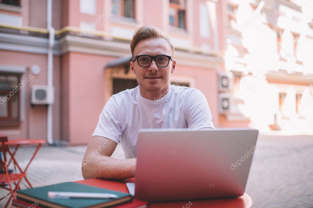 Portrait of skilled male IT professional in stylish eyewear looking at camera during break from online programming and initializing data system, successful man spending time for creating article