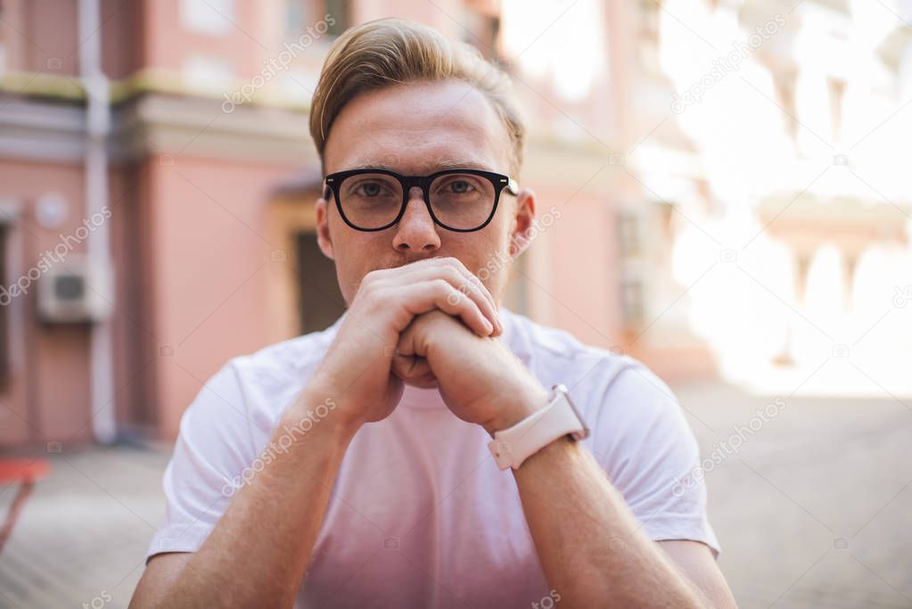 Close up portrait of handsome hipster guy in optical spectacles for vision correction looking at camera during leisure time outdoors, serious man in classic glasses sitting at urban setting
