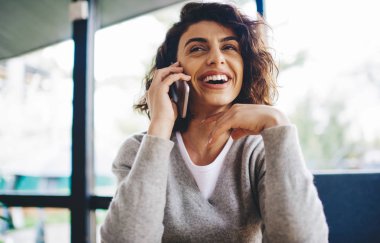 Birlikte çalışan kıvırcık saçlı neşeli genç bir kadın ve akıllı telefon cihazıyla iletişim kurarak boş zamanın tadını çıkarıyor. Hücresel olarak en iyi arkadaşıyla gülen pozitif hippi kız.