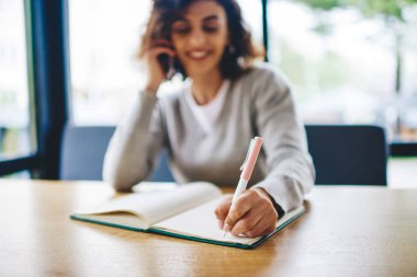Seçici bir şekilde elinde kalem tutan ve telefon görüşmesi sırasında not defterine not alan ahşap masada oturan öğrenciye odaklan. Bulanık öğrenci arıyor ve bilgi yazıyor.