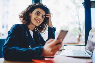 Akıllı telefonu elinde tutarken kameraya gülümseyen güzel hippi kız portresi ve kablosuz internet kullanarak dizüstü bilgisayarın başında çalışıyor. Bilgisayarda çalışan olumlu genç bir kadın.