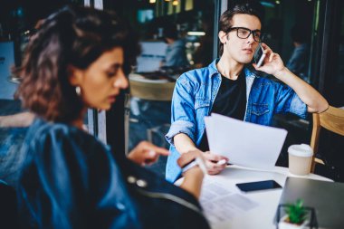 Genç işadamı akıllı telefon cihazını arıyor ve kağıt tutarak sorunları çözüyor ve modern uzayda masa başında bayan meslektaşla takım çalışması yapıyor. Hücresel konuşan hipster adama odaklanın.