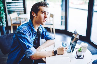 Bluetooth kulaklıklı düşünceli erkek öğrenci, modern teknoloji hakkında e-öğrenim için faydalı tezler yapmak için ders kitabı kullanarak, eğitim sesli kitabından edinilen bilgiler üzerinde yoğunlaştı ve kafa yordu.