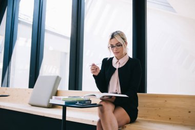 Modern iç mekan ile atölyede otururken, bilgi kitabından finansal verileri kontrol eden şık gözlüklü güzel bir yönetici, kapalı alandaki bilgiyi analiz eden başarılı bir yönetici.