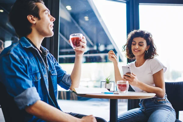 Cheerful female blogger messaging in online chat with internet followers on mobile phone device and using free 4G connection while ignoring live communication with best friend during meeting in cafe