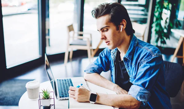 Intelligent male student writing essay for university while listening classic music for favourite playlist using bluetooth earphones and laptop application, millennial man doing homework indoors