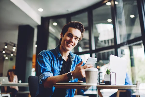 Portrait of cheerful caucasian young man smiling at camera while holding smartphone in hands and updating profile of blog sitting at laptop, generation of millennial guy using modern devices