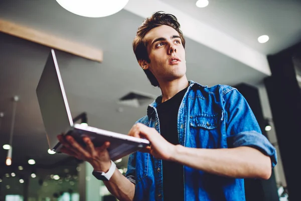 Contemplative skilled hipster guy pondering on received email from internet provider about connection on laptop computer, below view of pensive man in casual wear holding modern netbook indoors