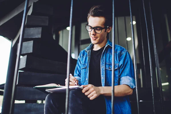 Positive hipster guy in eyewear for vision correction writing essay with good idea in notebook on leisure, successful hipster guy preparing to university exam and notes information for learning