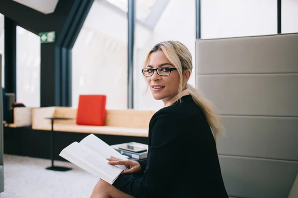 Portrait of beautiful female director in stylish elegant wear looking at camera while sitting in workshop with interesting literature book in hands, businesswoman in eyewear for vision correction