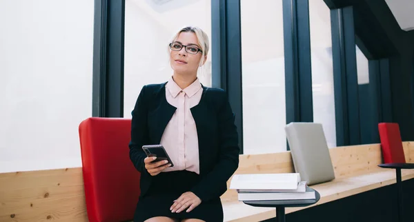有自信的女企业主穿着端庄雅致的西装坐在办公室里 戴着时髦眼镜 手持现代手机 看着室内相机的女性形象 — 图库照片
