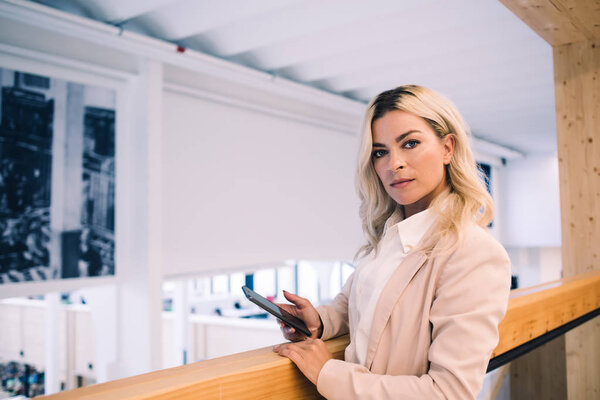 Женщина-директор в ожидании банковской операции на гаджете смартфона, портрет уверенной женщины с мобильным телефоном в руке, смотрящей в камеру в рабочее время в офисном здании