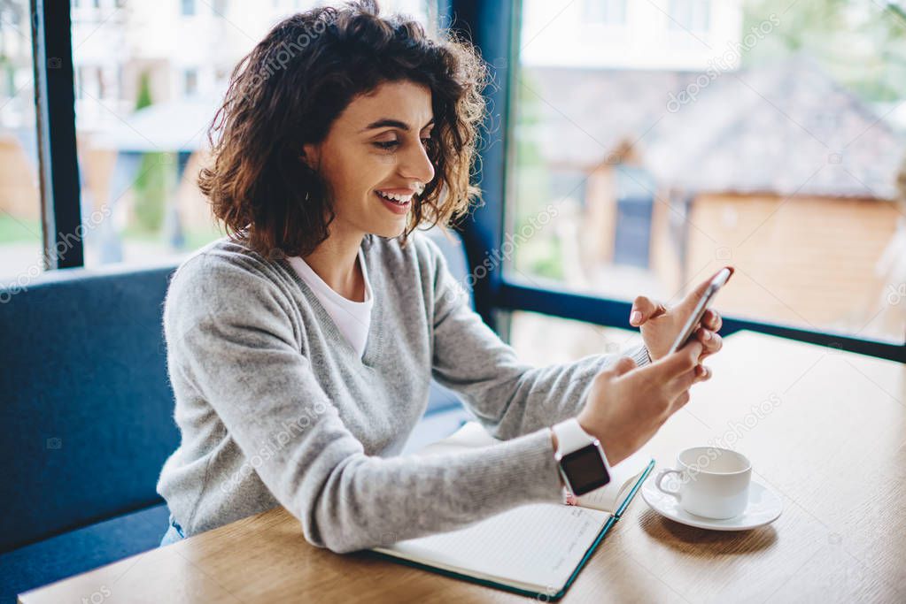 Cheerful young woman holding smartphone and watching video in social networks using 4G internet in coworking space.Positive caucasian blogger checking email on mobile phone sitting at desktop