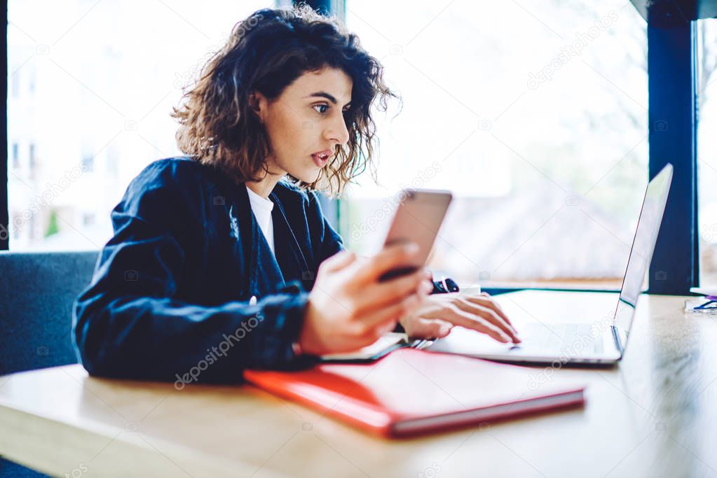 Young woman with short curly hair dressed in casual wear holding digital smartphone in hand while watching video on website using wireless internet on laptop computer in modern coworking space