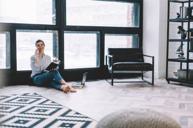 Dijital akıllı telefon cihazıyla arayan ve arkadaşlarıyla buluşmayı planladığı için konuşan, modern apartman dairesinde şık iç mimari ve büyük pencereli not defteriyle oturan genç bir kadın.